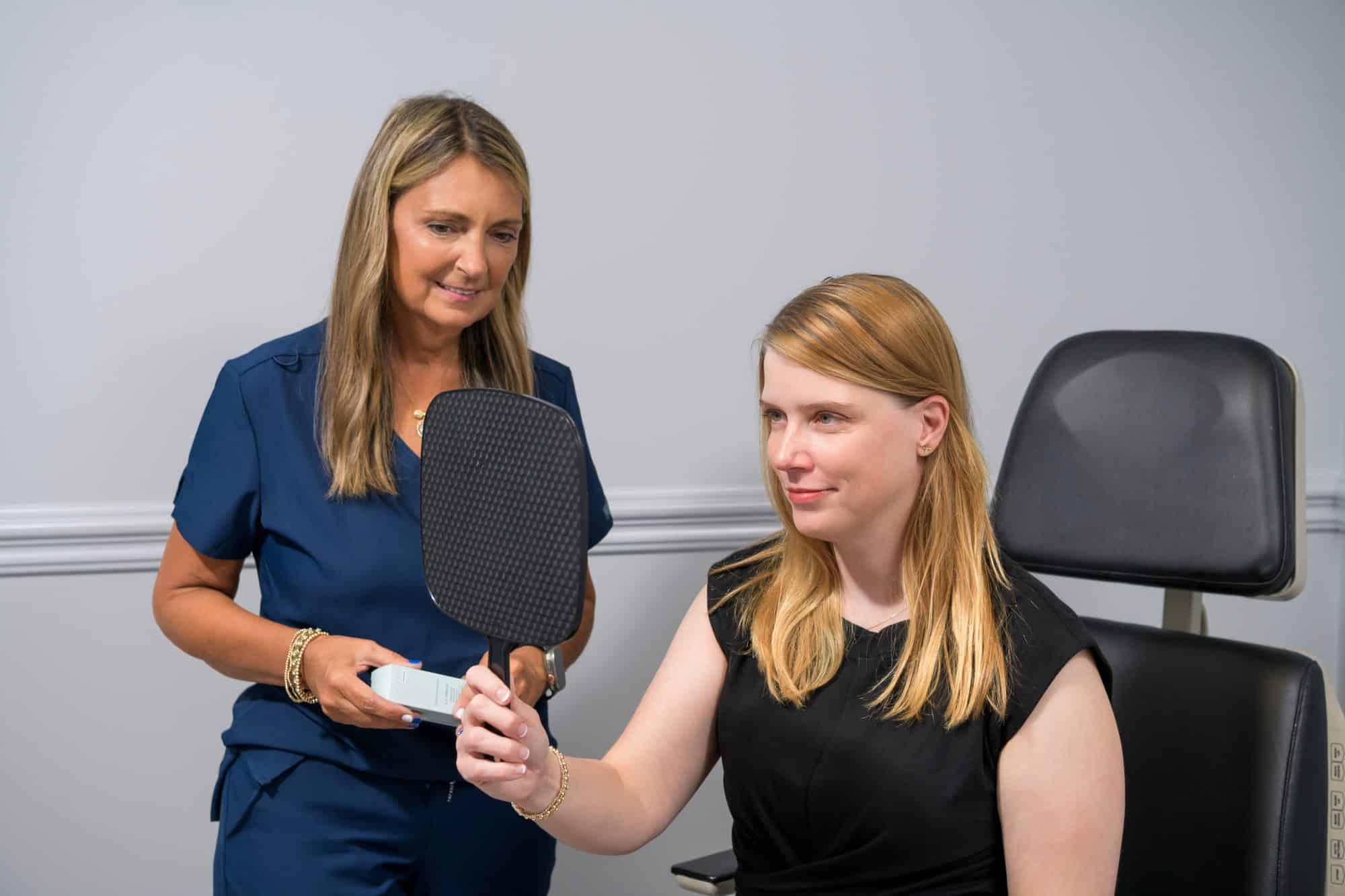 Sharon Simpson showing SkinCeuticals product to a caucasian female patient who's looking at her face in a handheld mirror